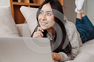 Joyful asian woman smiling and using laptop while lying on couch