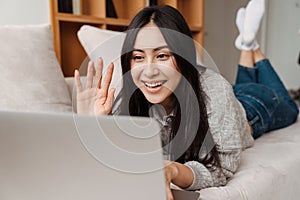 Joyful asian woman waving hand and using laptop while lying on couch