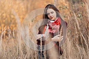 Joyful attractive fashionable young woman in a trendy knitted burgundy sweater with a stylish plaid scarf posing in a field among