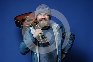 Joyful bearded man with an acoustic guitar, in a knitted hat and a waterproof jacket, gathered on a travel. Blue