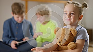 Joyful beautiful Caucasian little girl waving looking at camera sitting with teddy bear in living room as blurred boys