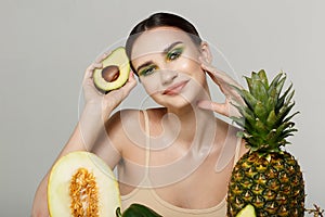 Joyful beautiful girl with bright art makeup holding sliced fresh avocado in hand near face