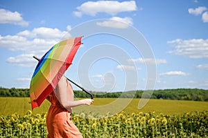 Joyful beautiful girl holding multicolored umbrella in sunflower field and blue cloud sky background