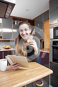 Joyful beautiful young woman holding a digital tablet in kitchen