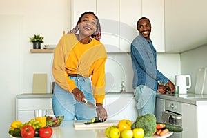 Joyful black couple cooking food and laughing, woman preparing salad for dinner, standing in modern kitchen