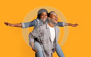 Joyful Black Couple Having Fun Together Over Yellow Background, Woman Piggybacking Boyfriend