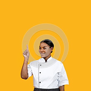Joyful black female chef with bright smile pointing upwards, suggesting successful idea