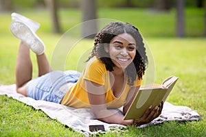 Joyful black girl reading interesting book while lying on picnic blanket at park