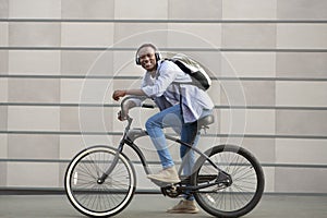 Joyful black guy with backpack and headphones riding bicycle near brick wall on city street