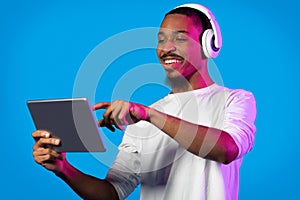Joyful black guy using modern digital pad and wireless headphones