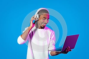 Joyful black guy using modern laptop and wireless headphones, telecommunication