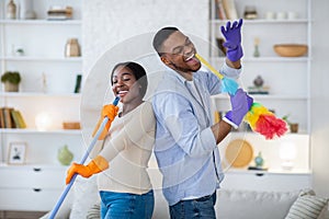 Joyful black man and his girlfriend singing during housecleaning, using mop and duster as microphones