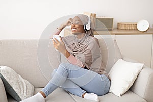 Joyful black Muslim woman in hijab and headphones listening to music on cellphone, closing her eyes in delight, at home