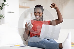 Joyful Black Woman Celebrating Success With Smartphone And Using Laptop At Home