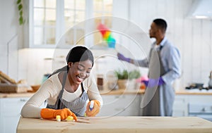 Joyful black woman cleaning kitchen table with detergent while her boyfriend dusting on background, empty space