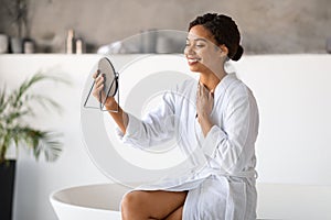 Joyful black woman in white bathrobe admiring her reflection in handheld mirror