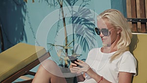 Joyful blonde girl checking mail in social networks while waiting friends near the hotel to swim in pool together