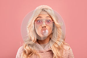 Joyful blonde lady chewing gum and blowing bubble, looking at camera on pink studio background