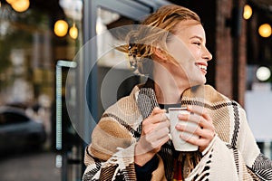 Joyful blonde woman in warm plaid drinking hot tea while stanging outdoors photo