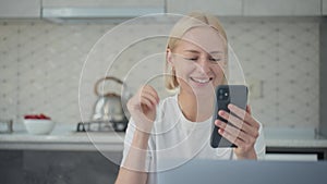 Joyful blonde woman waves bye in video call when sitting at laptop in kitchen