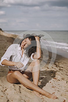 Joyful boho girl in white shirt sitting on sunny beach. Carefree stylish woman smiling and relaxing on seashore. Summer vacation.