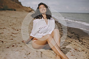 Joyful boho girl in white shirt sitting on sunny beach. Carefree stylish woman smiling and relaxing on seashore. Summer vacation.