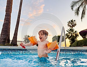 Joyful boy in arm bands enjoying sunset swimming pool