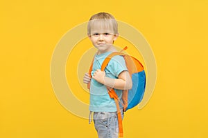 Joyful boy child stands with a backpack on his back and looks at the camera on a bright yellow background