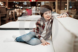 Joyful boy in store sits on bed in orthopedic mattress store. Check for softness of orthopedic furniture.