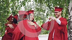 Joyful boys and girls graduates in gowns and hats are hugging congratulating each other on graduation, laughing and