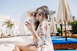 Joyful brunette girl checking mail in social neworks while waiting friends near the hotel to swim in pool together