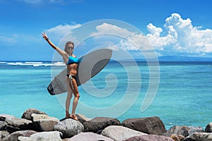 Joyful brunette woman stands on the rocks on the background of the sea and cloudy sky. She wears blue bikini, sunglasses
