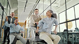 Joyful business team celebrating success. Four crazy happy colleagues riding chairs in office corridor, laughing