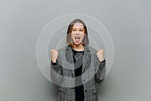 Joyful business woman in a jacket rejoices on a gray background with shouts and raised hands looking to the side, wearing smart