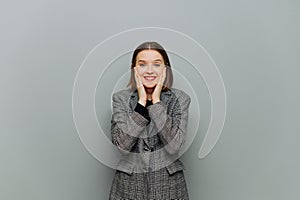 Joyful business woman in a jacket stands against the gray wall and is happy with a smile on her face looking at the camera