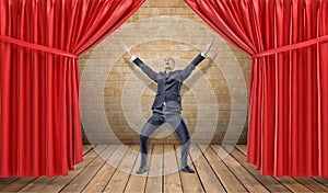 A joyful businessman standing at a wooden stage between red curtains in a victory pose.