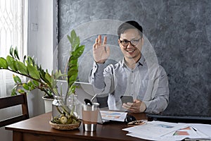Joyful Businessman Waving Hello Smiling To Camera Sitting In Modern Office. Entrepreneur Man Communicating Online Gesturing Hi