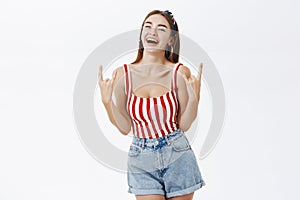 Joyful carefree stylish pinup girlfriend in striped top and denim shorts showing rock n roll gesture laughing amused