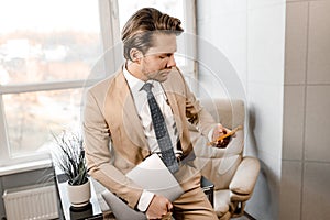 Joyful Caucasian businessman using smart phone standing in modern office hold laptop, male professional holding