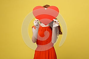 Joyful caucasian girl in orange gorgeous dress holding red heart cover her face. Shallow depth of field. Valentine day