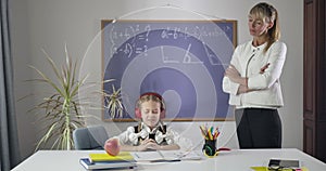 Joyful Caucasian girl sitting at the table in headphones and shaking head as adult tutor standing next to her