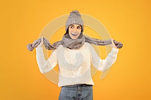 Joyful caucasian woman in warm sweater, grey knitted hat and scarf, posing and looking aside over yellow background