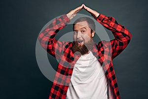 Joyful charming bearded man in red checkered shirt, raise hands above head and making roof or house gesture, smiling amused, have