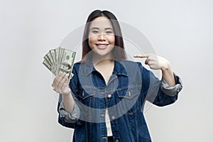 Joyful cheerful smiling woman in blue denim jacket standing holding and pointing ar dollar banknotes