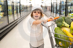 Joyful child boy in supermarket buys vegetables. Healthy food for children. Funny shopping. Cute kid with smile. Boy