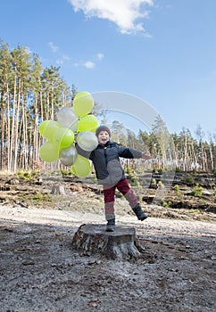 Joyful child preschooler boy holding balloons in his hands standing in the forest on a tree stump