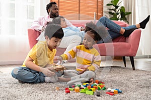 Joyful children sitting on carpet floor and playing with colorful wooden building blocks while parents relaxing on couch with