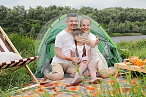 Joyful couple with kid on picnic, family sitting near tent on the ground, enjoying weekend together, spending happy time in open