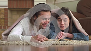 Joyful couple in love lying on the carpet in the bedroom covered with a blanket reading a book and cheerfully discusses