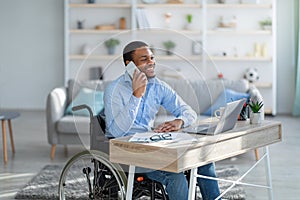 Joyful disabled black guy in wheelchair having phone conversation while working online on laptop from home